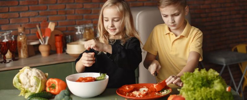 Serious boy cutting pepper for vegetable salad while standing near happy sister seasoning ingredients put into deep bowl in loft style kitchen