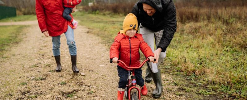 Happy family walking outdoors in fall, teaching child to ride bike.